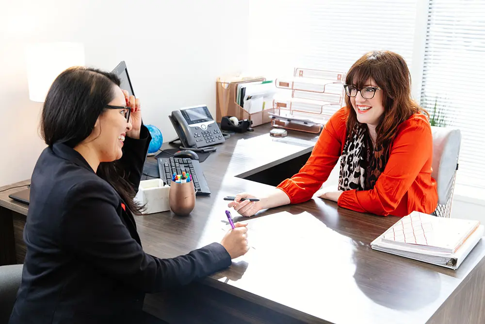 Two women are seated at a modern office desk, engaged in a pleasant conversation. One woman, in a black blazer, holds a pen over a document, while the other, in a red blouse with a black-and-white scarf, listens attentively with a smile.