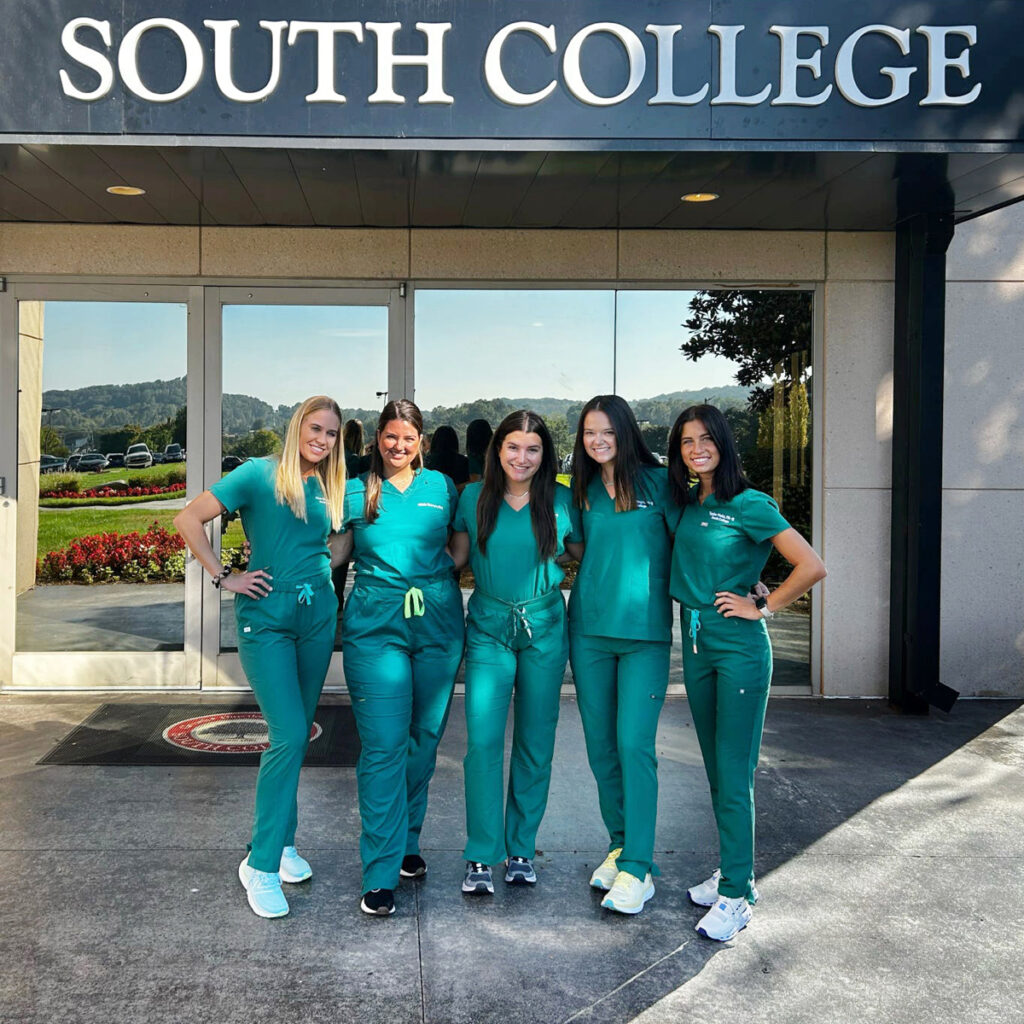 Five people in green medical scrubs stand together smiling in front of a building labeled "South College". They are outdoors, standing side by side with arms around each other. The surroundings include a glass door and the partial view of a garden.