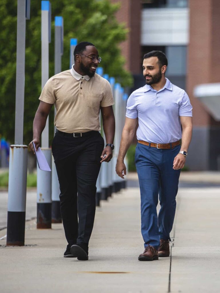 Two men are walking on a sidewalk, engaged in conversation. One is wearing a beige polo shirt and black pants, holding a paper. The other is wearing a light blue polo shirt and blue pants. Trees and a building are in the background.
