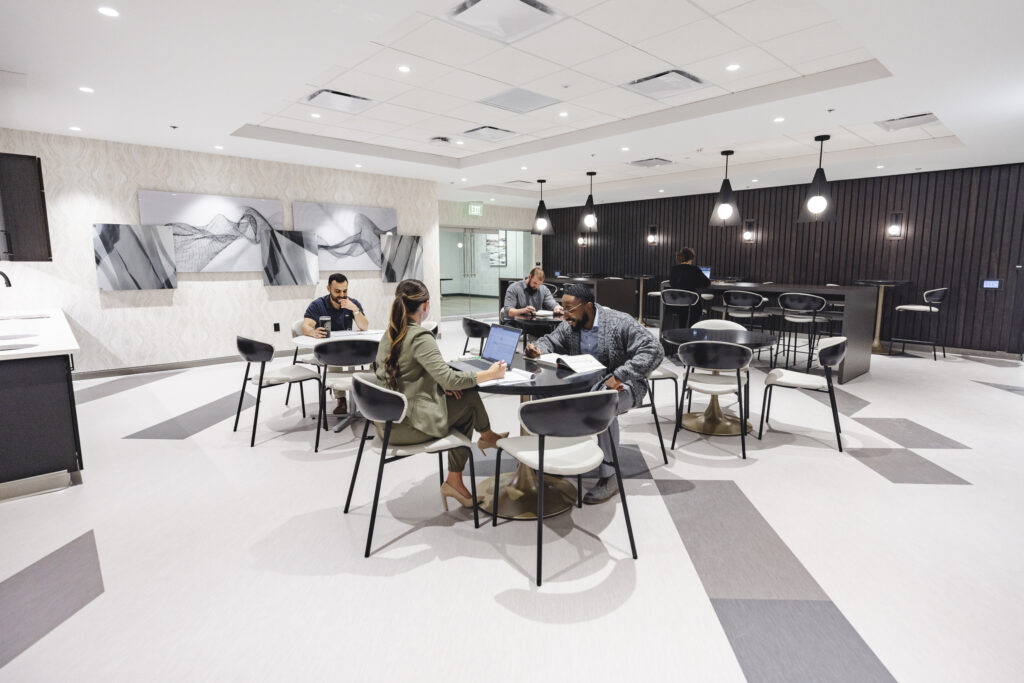 A modern, open-concept student lounge with white walls and ceiling, featuring several small tables and chairs. Four people are seated at different tables, using laptops and engaging in discussions. The space is well-lit with hanging pendant lights and has contemporary decor.