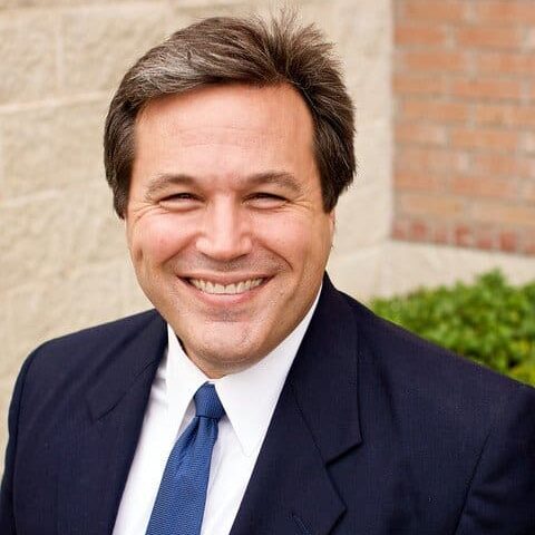 A man with a blue tie smiles at the camera. He appears to be outdoors.