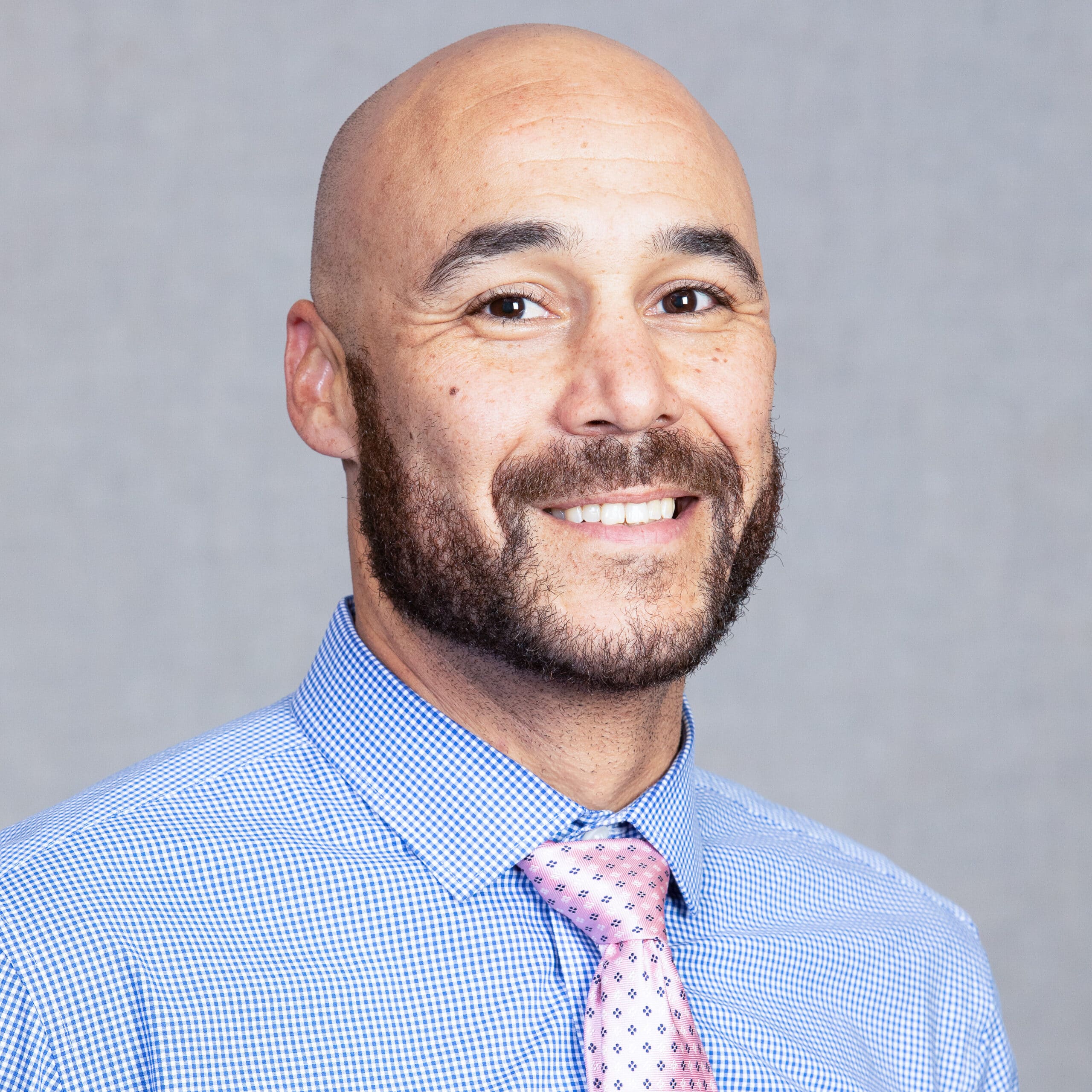 A man with a full beard is smiling. He is wearing a blue checkered shirt and a pink patterned tie.