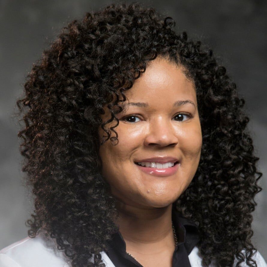 Smiling woman with curly hair, wearing a white blazer over a black top, posing against a dark gray background.