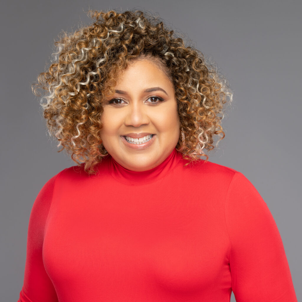 A woman with curly hair smiles at the camera, wearing a bright red turtleneck. The background is a plain gray.