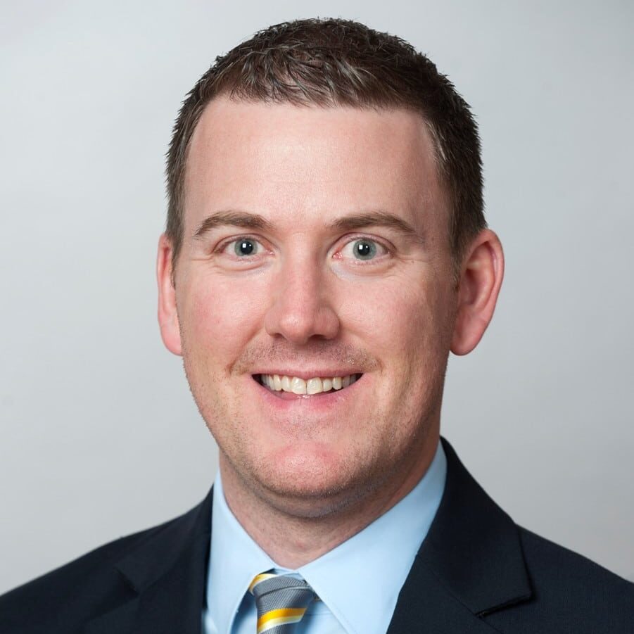 Man in a dark suit, light blue shirt, and striped tie smiling at the camera against a plain background.