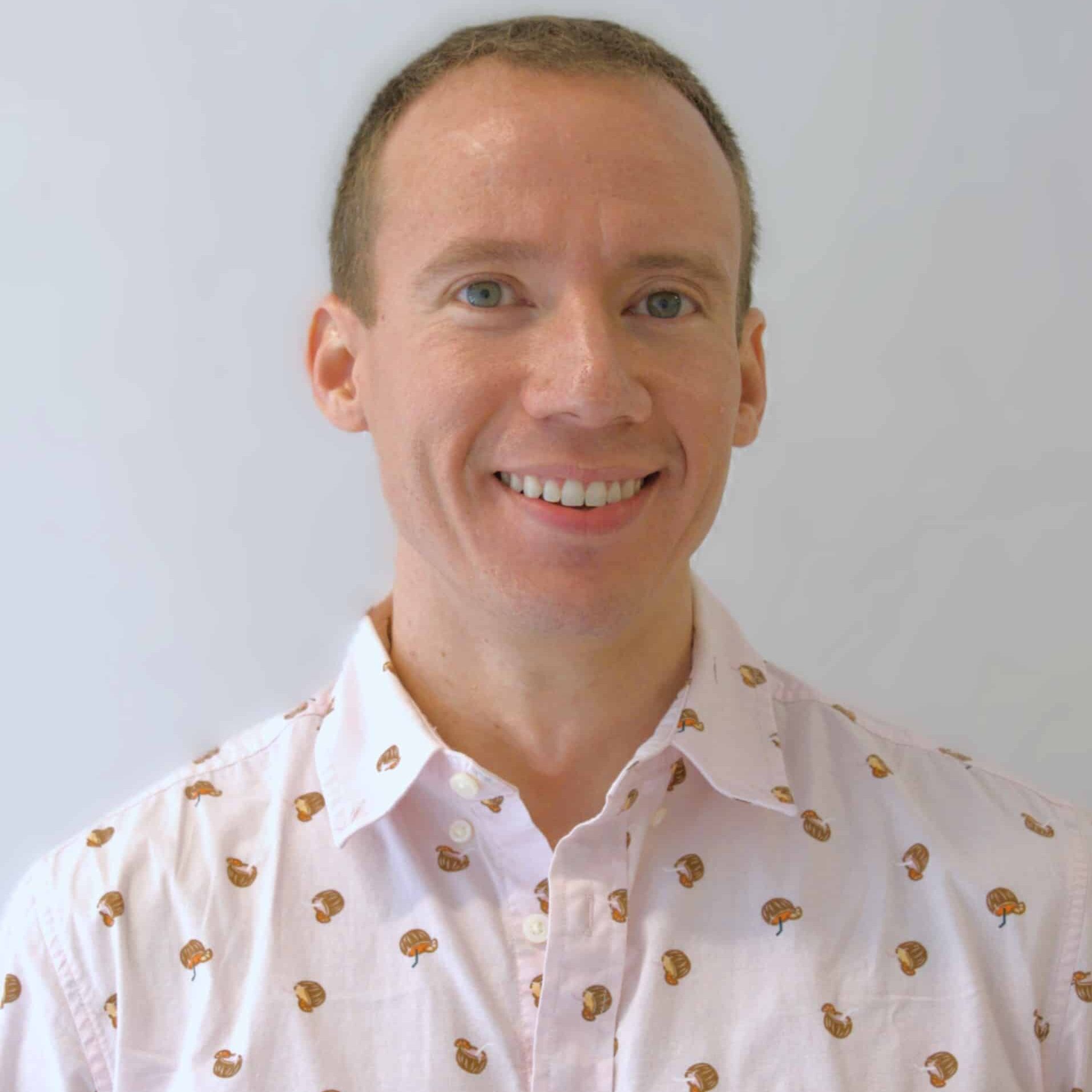 A person with short hair smiles while wearing a light pink shirt adorned with a pattern of small, colorful cupcakes against a plain white background.