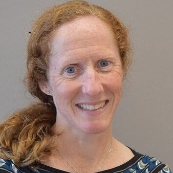 A woman with red hair and blue eyes smiles at the camera. She is wearing a patterned blue and black top.