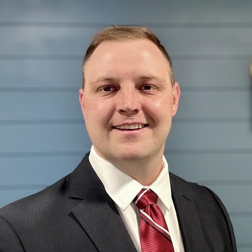 A person wearing a dark suit, white shirt, and red tie stands smiling against a light blue, horizontal panel wall.