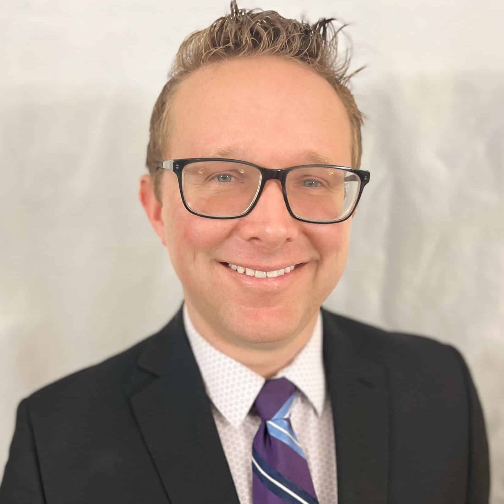 A person wearing glasses and a suit with a white shirt and striped blue tie is smiling.