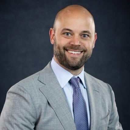 A smiling man with a beard is wearing a light gray suit, white shirt, and purple tie.