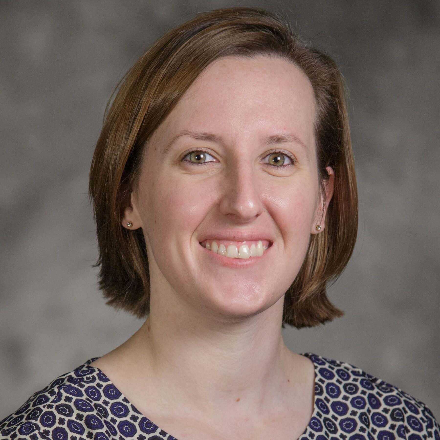 A person with short brown hair smiles at the camera, wearing a long-sleeved shirt with a blue and white geometric pattern.